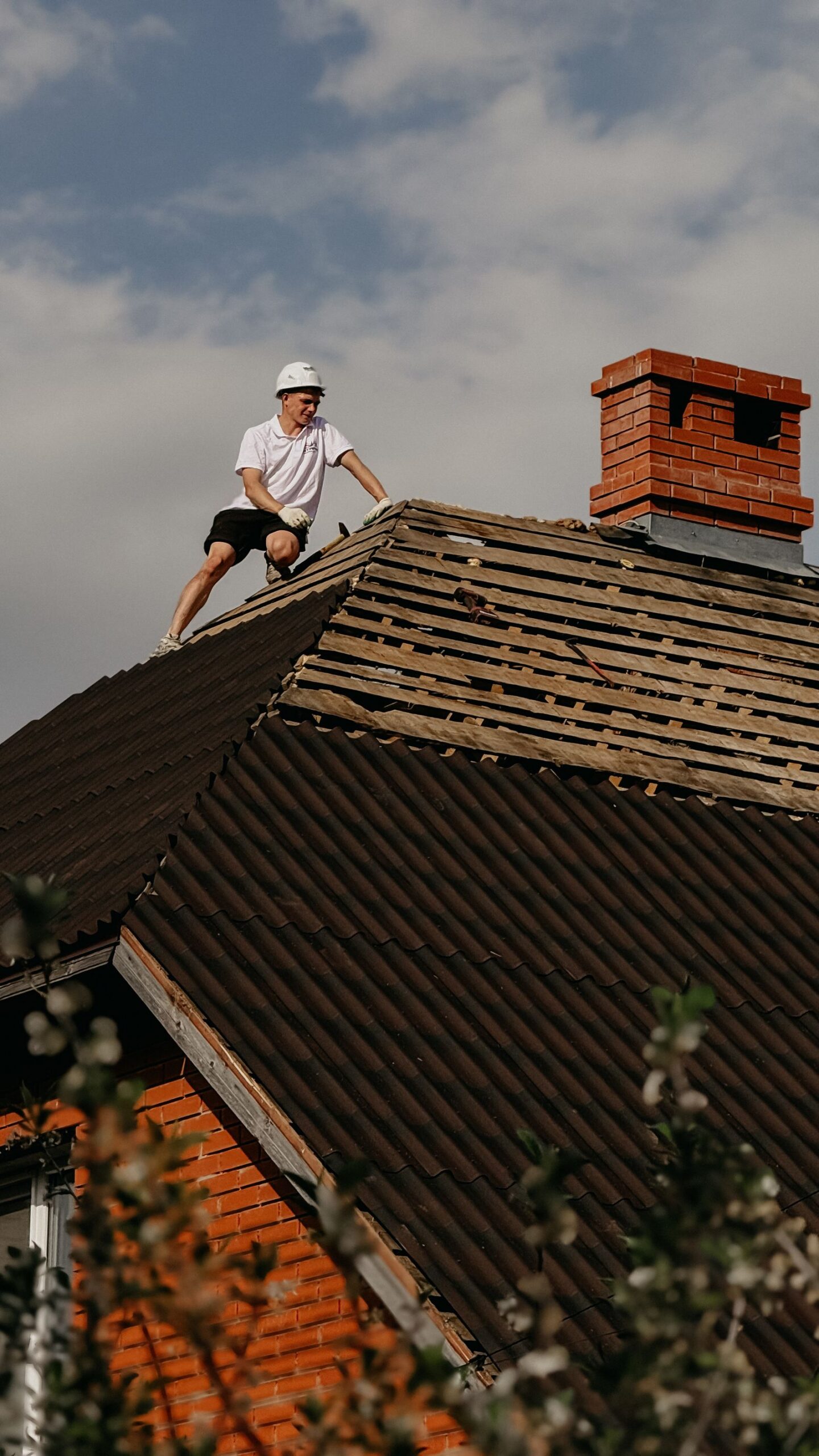 Roofer in a safety helmet repairing the shingle roof of a red brick house under a clear sky
